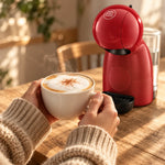Manos con un capuchino recién hecho junto a la cafetera Nescafé® Dolce Gusto® Piccolo XS roja