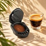 Cápsula recargable iCafilas abierta con café y un vaso de espresso bajo luz de sol entre hojas